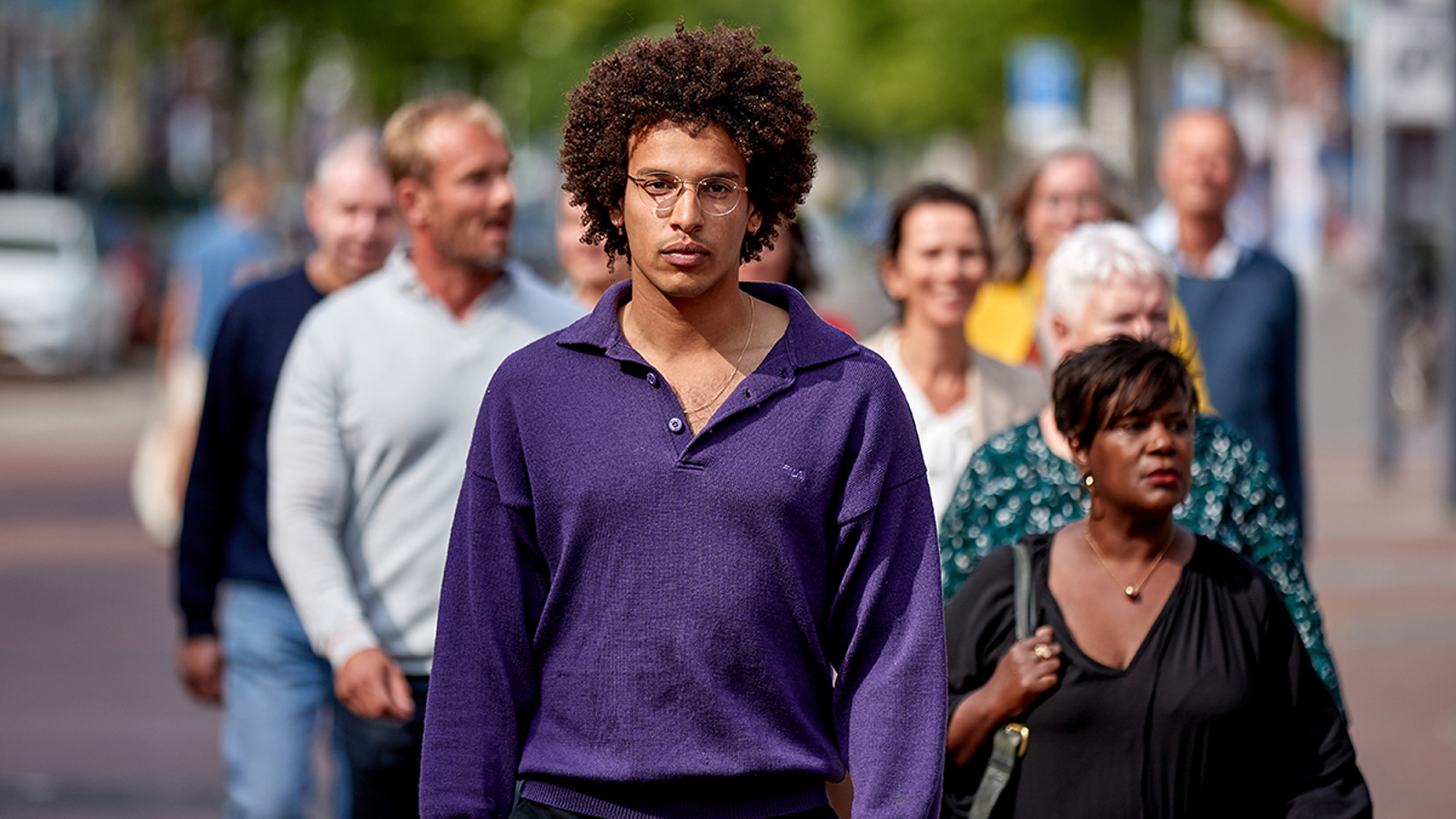 man met donkere krullen die stilstaat tussen een groep bewegende mensen en recht de camera inkijkt